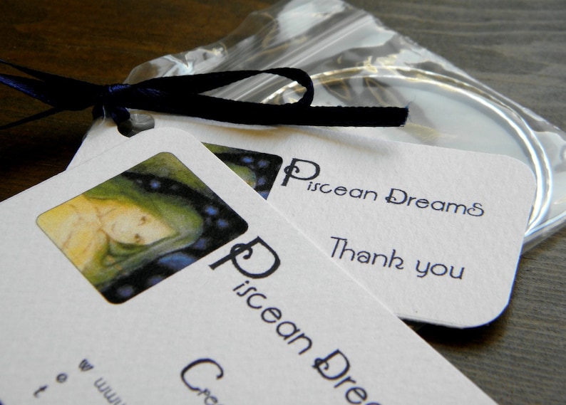 May include: Two white cards with the text "Piscean Dreams" and "Thank you" on them. The cards are on top of a clear plastic bag with a silver ring inside. The top card has a small illustration of a woman with blue and green hair.