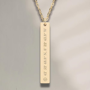May include: A gold-colored rectangular pendant necklace with engraved coordinates: 42° 52' 48.8" N, 8" 32' 43.8" W. The pendant hangs from a delicate chain. The necklace is displayed on a neutral background, suggesting a minimalist aesthetic.