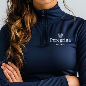 May include: Navy blue long-sleeve athletic top with a quarter-zip neckline. The top features the white text "Peregrina EST. 2025" and a seashell logo. The model is wearing the top. The word "Navy" is in a white box at the bottom.