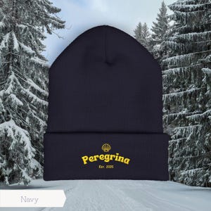 May include: Navy blue beanie with the word "Peregrina" embroidered in yellow, along with a seashell graphic and the text "Est. 2025". The beanie is set against a snowy winter backdrop.