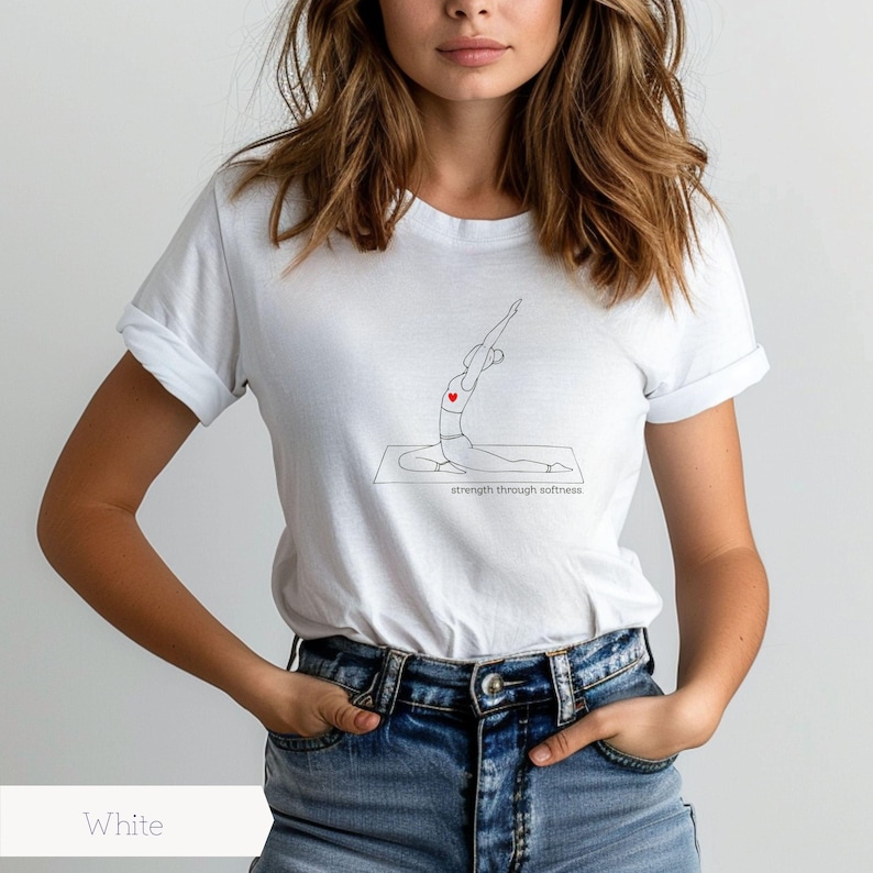 May include: White t-shirt featuring a minimalist line drawing of a person in a yoga pose with a red heart. The text "strength through softness" is below the drawing. The shirt is paired with blue jeans.