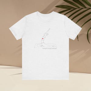 May include: A light gray t-shirt featuring a minimalist line drawing of a person in a yoga pose with a red heart. The text "strength through softness" is below the drawing. The shirt is made of a soft material.