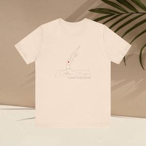 May include: Cream-colored t-shirt featuring a minimalist line drawing of a person in a yoga pose, with a red heart on the chest. The text "strength through softness" is printed below the illustration. The shirt has short sleeves and a crew neck.