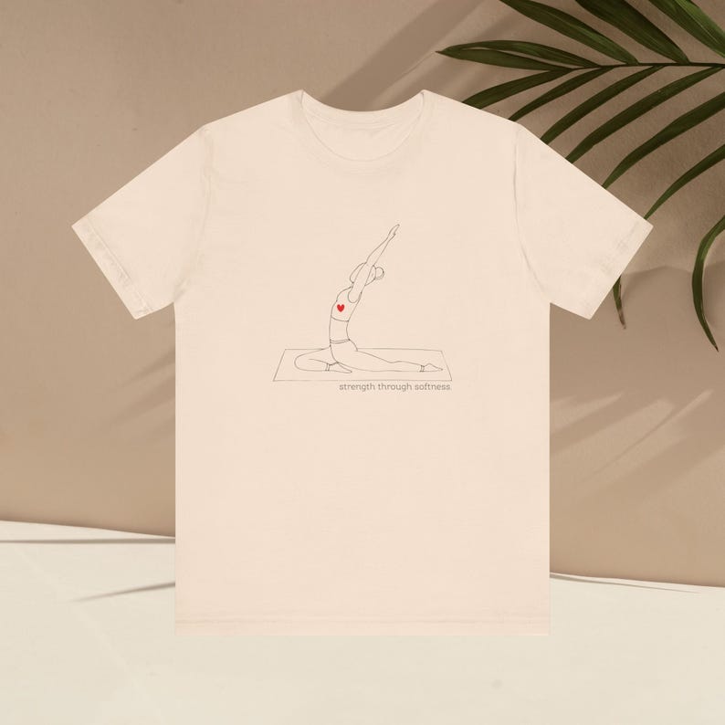 May include: Cream-colored t-shirt featuring a minimalist line drawing of a person in a yoga pose, with a red heart on the chest. The text "strength through softness" is printed below the illustration. The shirt has short sleeves and a crew neck.