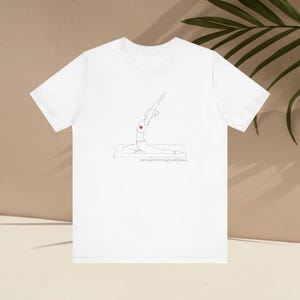 May include: White t-shirt featuring a minimalist line drawing of a person in a yoga pose, with a red heart and the text "strength through softness" in a thin, gray font. The shirt is made of a soft material.