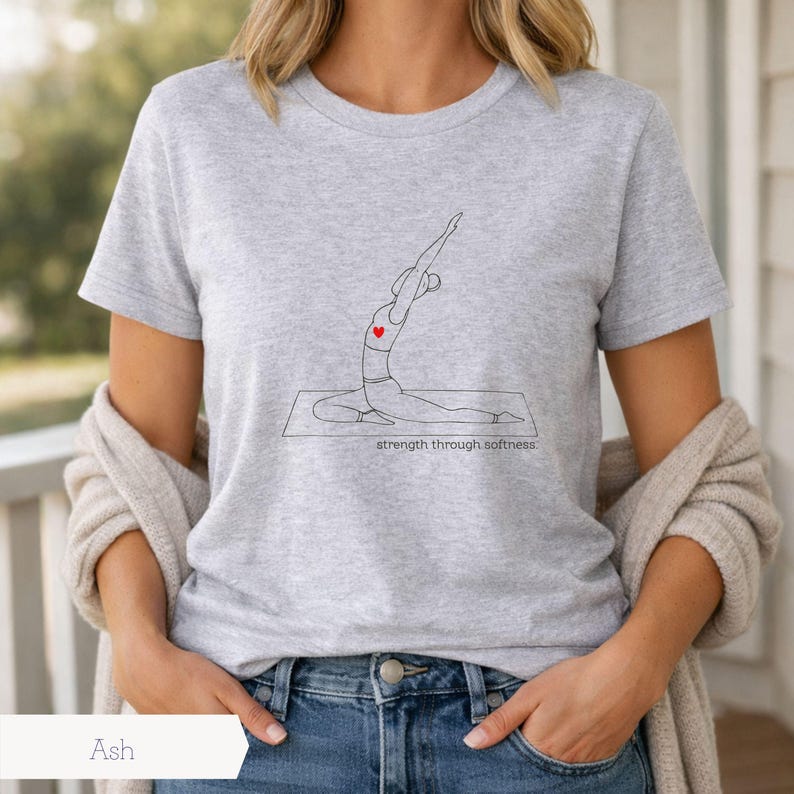 May include: Ash gray t-shirt featuring a line drawing of a person in a yoga pose with a red heart. The text "strength through softness" is below the drawing. The shirt is paired with blue jeans.