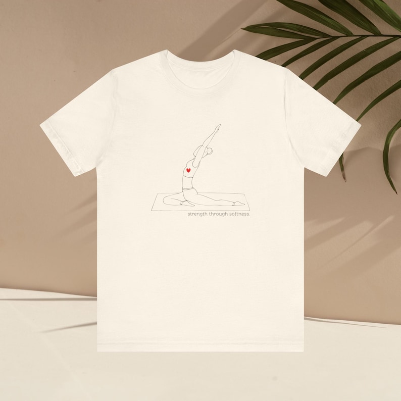 May include: Off-white t-shirt featuring a minimalist line drawing of a person in a yoga pose, with a red heart. The text "strength through softness" is below the drawing. The shirt is made of a soft material.