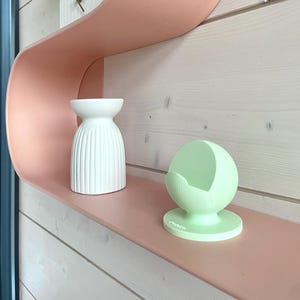 May include: A modern, wavy, peach-colored shelf displays a white, ribbed vase, a roll of white paper, and a light green spherical object. The shelf is mounted on a light wood-paneled wall, creating a minimalist aesthetic.