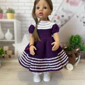 May include: A doll wearing a purple crochet dress with white accents. The dress features a white crocheted collar and a skirt with horizontal white stripes. The doll has brown hair and is wearing white shoes. The background includes a white brick wall and a decorative tent.
