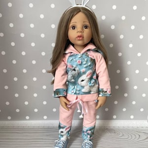 May include: A doll wearing a pink and blue bunny-themed outfit. The outfit includes a hooded sweatshirt with a bunny and floral design, matching pants, and blue sneakers. The doll also wears a white bunny ear headband.