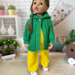 May include: A doll wearing a green knitted hooded jacket over a yellow jumpsuit. The doll is also wearing white sneakers with red accents. The doll has brown hair and is standing in front of a white brick wall.