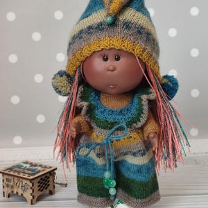 Könnte beinhalten: Eine handgefertigte Puppe in einem gestrickten Outfit in Blau-, Grün-, Gelb- und Brauntönen. Die Puppe hat lange, rosa-orangefarbene Haare und einen passenden Hut mit Bommeln. Die Puppe trägt weiße Schuhe und hat eine kleine hölzerne Spieluhr daneben.