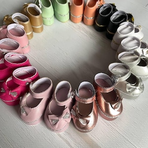 Könnte beinhalten: Eine kreisförmige Anordnung von Miniatur-Puppenschuhen in verschiedenen Farben, darunter Rosa, Gold, Grün und Schwarz. Jeder Schuh hat einen Riemen und eine Schleife mit silbernen Akzenten. Die Schuhe sind auf einer hellen Oberfläche angeordnet.
