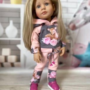 May include: A doll with long blonde hair wearing a pink and gray outfit. The outfit includes a hooded sweatshirt with a deer and floral design, matching pants, and pink shoes. The doll is standing in front of a white brick wall.