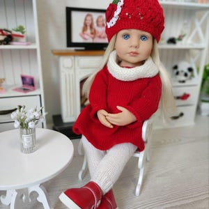 May include: A doll wearing a red knit dress, matching hat with a white floral accent, and red shoes with white soles. The doll has long blonde hair, blue eyes, and is wearing a white scarf and white knit leggings. The doll is sitting on a white chair.