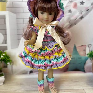 Könnte beinhalten: Eine Puppe in einem handgefertigten Häkel-Outfit. Das Outfit umfasst ein mehrfarbiges Kleid mit Rüschen, eine passende Haube und Stiefel. Das Kleid hat gelbe, rosa, lila und türkisfarbene Farben mit einer cremefarbenen Schleife um die Taille.