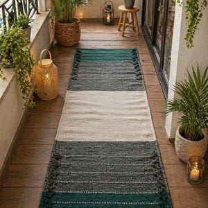Puede incluir: Una alfombra rectangular con un dise&ntilde;o de bloques de color en tonos verdes, grises y crema. La alfombra tiene un aspecto texturizado con flecos en los extremos cortos. La alfombra est&aacute; colocada sobre un suelo de madera, creando un ambiente acogedor.