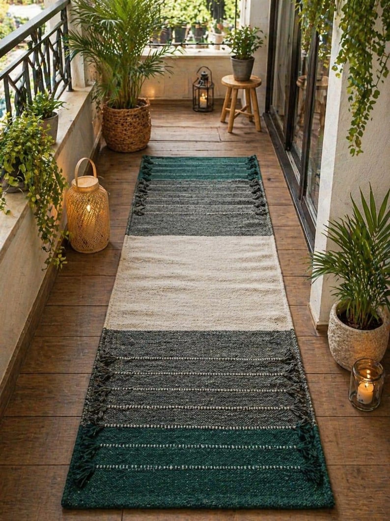 Puede incluir: Una alfombra rectangular con un dise&ntilde;o de bloques de color en tonos verdes, grises y crema. La alfombra tiene un aspecto texturizado con flecos en los extremos cortos. La alfombra est&aacute; colocada sobre un suelo de madera, creando un ambiente acogedor.