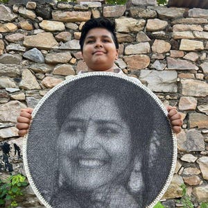 Può includere: Un ritratto rotondo in filo nero e bianco di una persona sorridente. L'opera d'arte è tenuta da una persona davanti a un muro di pietra. Il ritratto ha un bordo bianco.