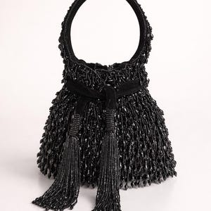 Könnte beinhalten: Eine schwarze Handtasche mit einer durchgehend perlenbesetzten Textur und einem kreisförmigen Griff. Die Tasche verfügt über einen Samt-Kordelzugverschluss an der Oberseite, akzentuiert durch zwei lange, herabhängende Perlenquasten.