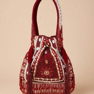 Może przedstawiać: Czerwona torebka z koralikami, z aksamitną rączką i frędzlami z koralików. Torba ma kwiatowy wzór z białymi i złotymi koralikami. Rączka ma wysokość około 30 cm.