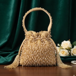 May include: A gold beaded handbag with a rounded handle and drawstring closure. The bag features long tassels and is adorned with small, dangling beads. The background includes white roses and a dark green velvet curtain.