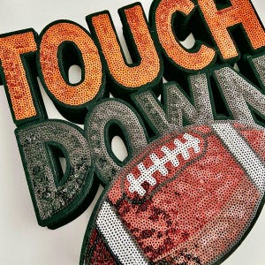 May include: A decorative piece spelling "TOUCH DOWN" in orange and silver sequins, with a sequined football. The letters have a dark green border, and the football is red and white.