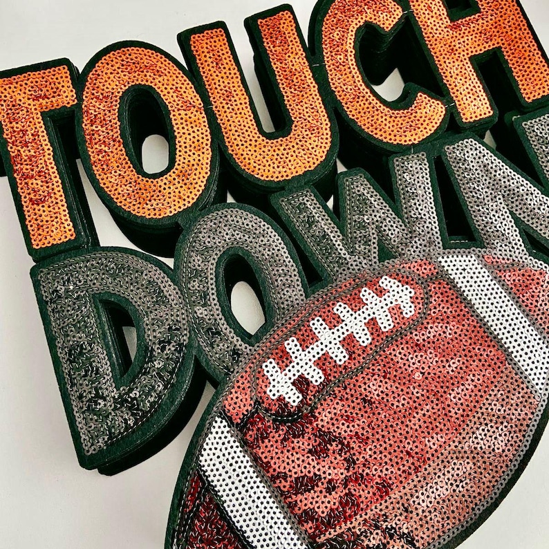 May include: A decorative piece spelling "TOUCH DOWN" in orange and silver sequins, with a sequined football. The letters have a dark green border, and the football is red and white.