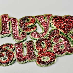 May include: Sequined letters spelling "Hey Suga" in red and pink sequins with a green glitter outline. A sequined heart shape is also present. The letters are arranged on a white surface.