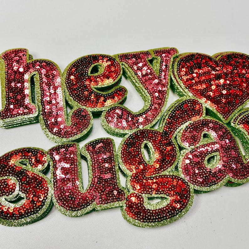May include: Sequined letters spelling "Hey Suga" in red and pink sequins with a green glitter outline. A sequined heart shape is also present. The letters are arranged on a white surface.