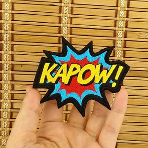 K&ouml;nnte beinhalten: Ein farbenfroher Aufn&auml;her mit dem Wort "KAPOW!" in Gelb, umrandet von Schwarz, vor einem roten und blauen Explosionsdesign. Der Aufn&auml;her ist wahrscheinlich f&uuml;r Kleidung oder Accessoires.