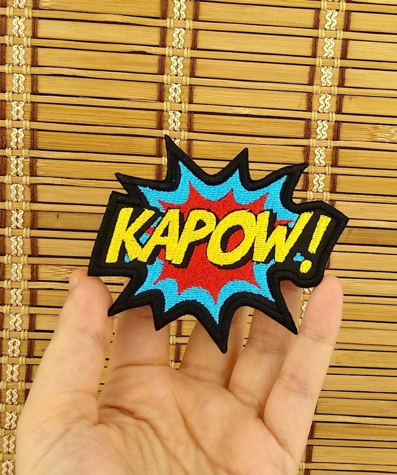 K&ouml;nnte beinhalten: Ein farbenfroher Aufn&auml;her mit dem Wort "KAPOW!" in Gelb, umrandet von Schwarz, vor einem roten und blauen Explosionsdesign. Der Aufn&auml;her ist wahrscheinlich f&uuml;r Kleidung oder Accessoires.