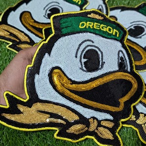 May include: Embroidered patches of the Oregon Ducks mascot. The patches have a light blue background, black outlines, and gold sequin accents. The word "OREGON" is embroidered in gold on a green hat.