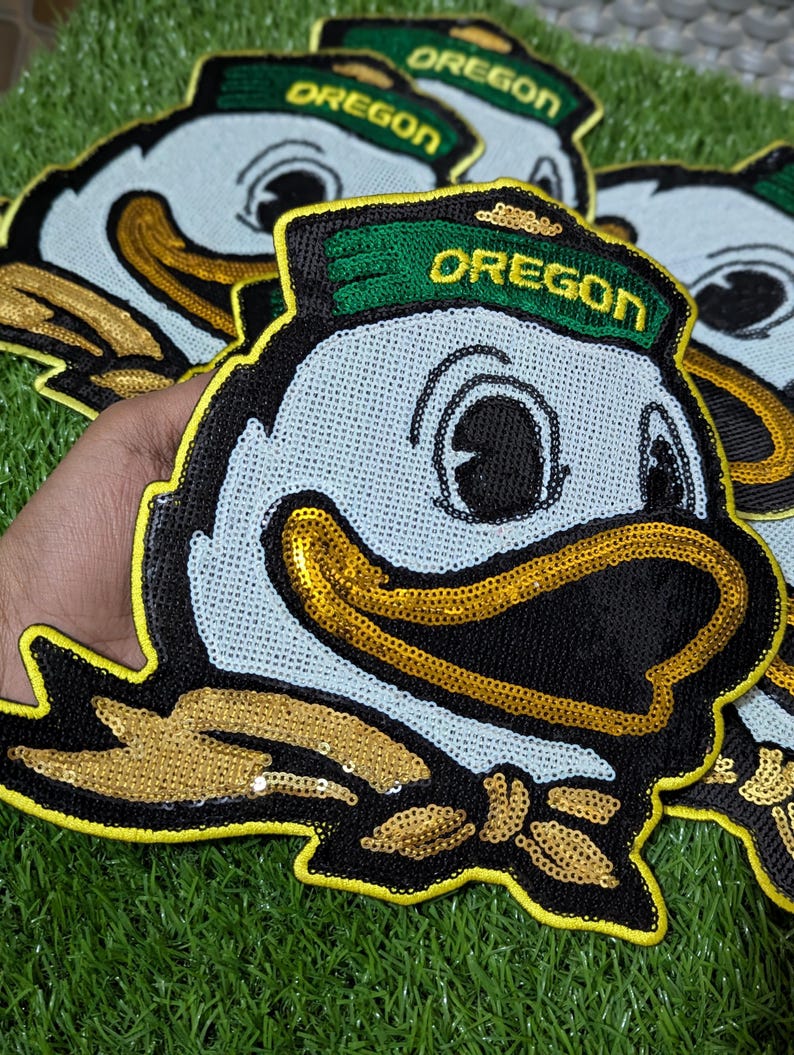 May include: Embroidered patches of the Oregon Ducks mascot. The patches have a light blue background, black outlines, and gold sequin accents. The word "OREGON" is embroidered in gold on a green hat.