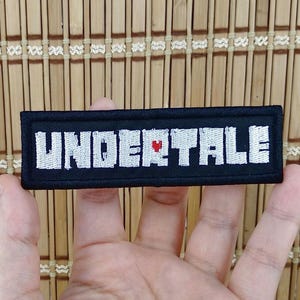 K&ouml;nnte beinhalten: Ein schwarzer rechteckiger Aufn&auml;her mit dem Wort "UNDERTALE" in wei&szlig;en Gro&szlig;buchstaben gestickt. Ein kleines rotes Herz ersetzt den Buchstaben "E" in der Mitte des Wortes. Der Aufn&auml;her wird in einer Hand gehalten.