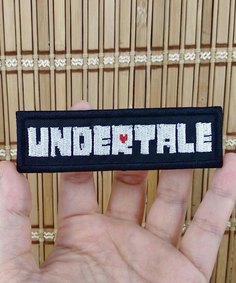 K&ouml;nnte beinhalten: Ein schwarzer rechteckiger Aufn&auml;her mit dem Wort "UNDERTALE" in wei&szlig;en Gro&szlig;buchstaben gestickt. Ein kleines rotes Herz ersetzt den Buchstaben "E" in der Mitte des Wortes. Der Aufn&auml;her wird in einer Hand gehalten.
