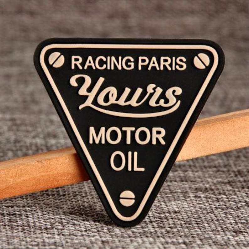 May include: Black triangular patch with white trim and text. The text reads "RACING PARIS Yours MOTOR OIL". The patch is set against a textured gray background and a wooden stick.