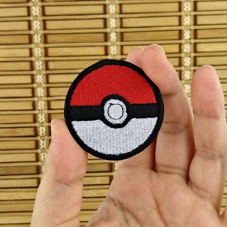 K&ouml;nnte beinhalten: Runder, gestickter Aufn&auml;her mit rotem und wei&szlig;em Design und schwarzem Rand. Der Aufn&auml;her &auml;hnelt einem Cartoon-Ball. Der Aufn&auml;her wird in einer Hand vor einem Bambushintergrund gehalten.