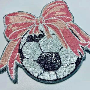 May include: A sequined soccer ball design with a large pink bow. The soccer ball is silver and black, and the bow is pink and peach. The design is outlined in silver glitter.