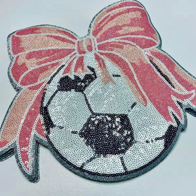 May include: A sequined soccer ball design with a large pink bow. The soccer ball is silver and black, and the bow is pink and peach. The design is outlined in silver glitter.