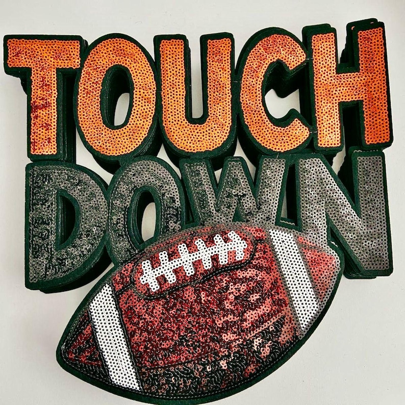 May include: A decorative piece with the words "TOUCH DOWN" in orange and silver sequins. A football-shaped object with red and white sequins is in the foreground. The letters have a dark green border.
