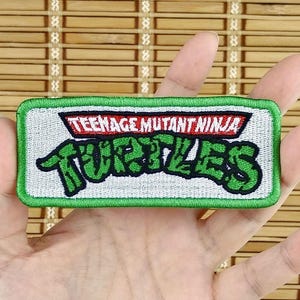 K&ouml;nnte beinhalten: Ein rechteckiger Aufn&auml;her mit dem Schriftzug "TEENAGE MUTANT NINJA TURTLES" in gr&uuml;nen und roten Buchstaben. Der Aufn&auml;her hat einen wei&szlig;en Hintergrund und einen gr&uuml;nen Rand. Der Aufn&auml;her wird in der Hand einer Person gehalten.