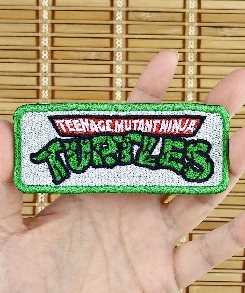 K&ouml;nnte beinhalten: Ein rechteckiger Aufn&auml;her mit dem Schriftzug "TEENAGE MUTANT NINJA TURTLES" in gr&uuml;nen und roten Buchstaben. Der Aufn&auml;her hat einen wei&szlig;en Hintergrund und einen gr&uuml;nen Rand. Der Aufn&auml;her wird in der Hand einer Person gehalten.
