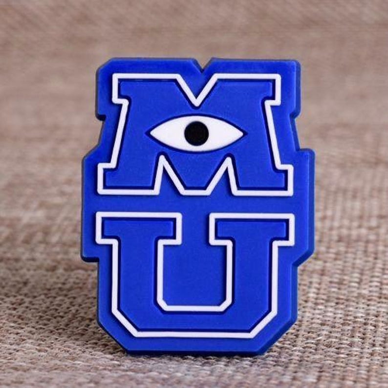 May include: A blue and white emblem featuring the letters "M" and "U" with an eye in the center. The design is outlined in white, creating a bold contrast against the blue background. The emblem is likely a decorative item.