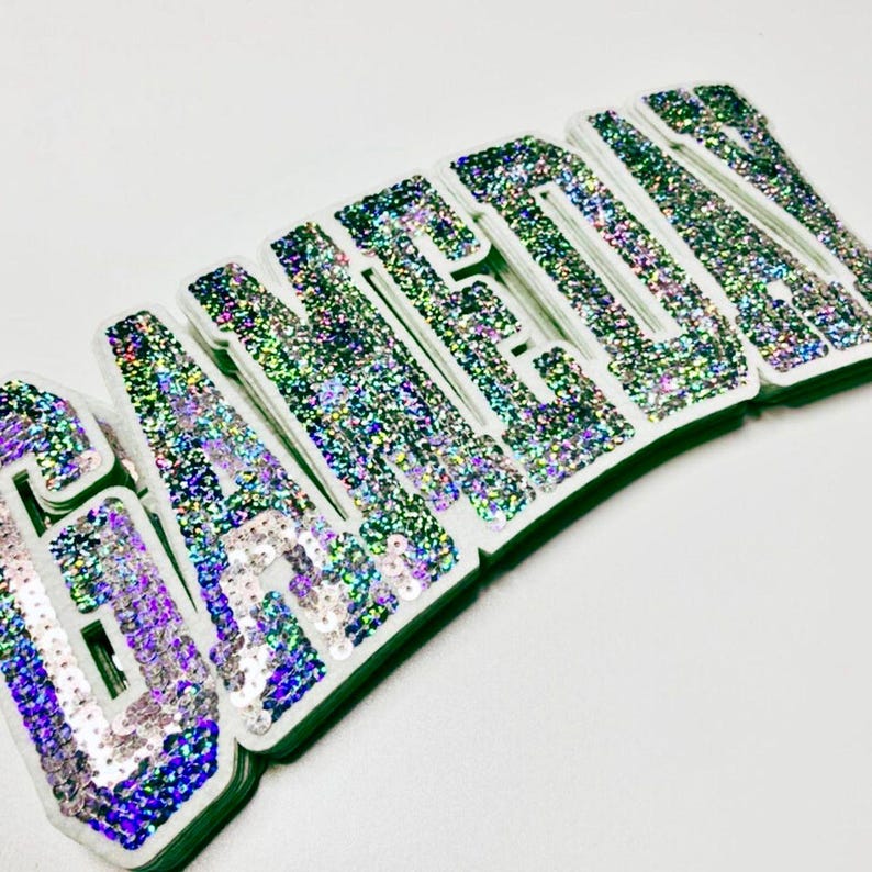 May include: Close-up of the word "GANEDEN" in large, glittery, sequined letters. The letters are white with iridescent sequins and a green border. The letters are stacked on top of each other.