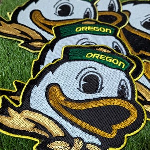 May include: Embroidered patches featuring the Oregon Ducks mascot. The patches have a light blue face, a black beak, and a green hat with the word "OREGON" in gold. The patches are outlined in yellow and gold sequins.