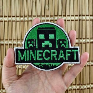 K&ouml;nnte beinhalten: Ein Minecraft-Aufn&auml;her mit gr&uuml;nem und schwarzem Design. Der Aufn&auml;her zeigt das Wort "MINECRAFT" in wei&szlig;en Buchstaben auf schwarzem Hintergrund. Der Aufn&auml;her hat einen gr&uuml;nen kreisf&ouml;rmigen Rand mit einem schwarzen pixeligen Charakter.