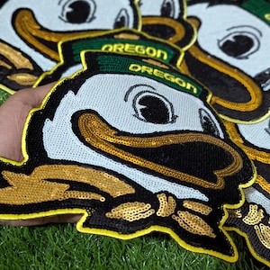 May include: A selection of sequined Oregon Duck patches. The patches feature a white face, black beak, and gold sequin detailing. The text "OREGON" is on a green hat.