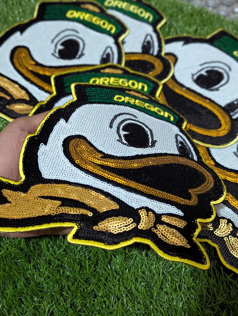 May include: A selection of sequined Oregon Duck patches. The patches feature a white face, black beak, and gold sequin detailing. The text "OREGON" is on a green hat.
