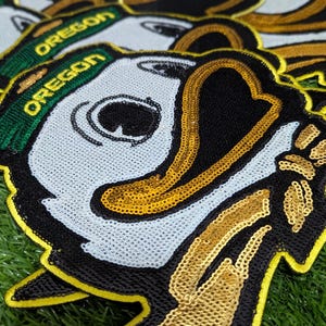 May include: Close-up of several sequined patches depicting the Oregon Ducks mascot. The patches have a white face, black beak, and a green and gold "OREGON" banner. The patches are on a green surface.